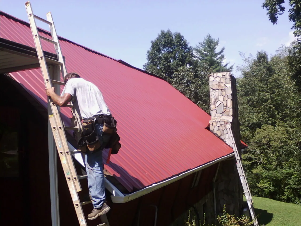 Roofer on a ladder beside a red metal roof and stone chimney — Soffit & Fascia Repair by Roofing Brothers in Sublime